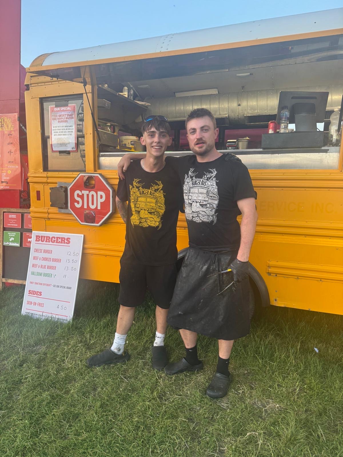Two Burger Bus crew members arm in arm by the bus and its menu board